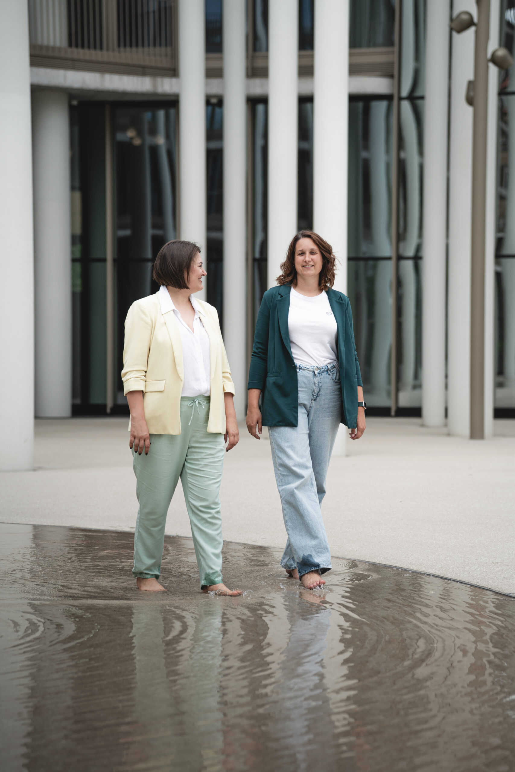 Zwei Frauen in Blazern laufen barfuß durch ein flaches Wasserbecken vor moderner Architektur.